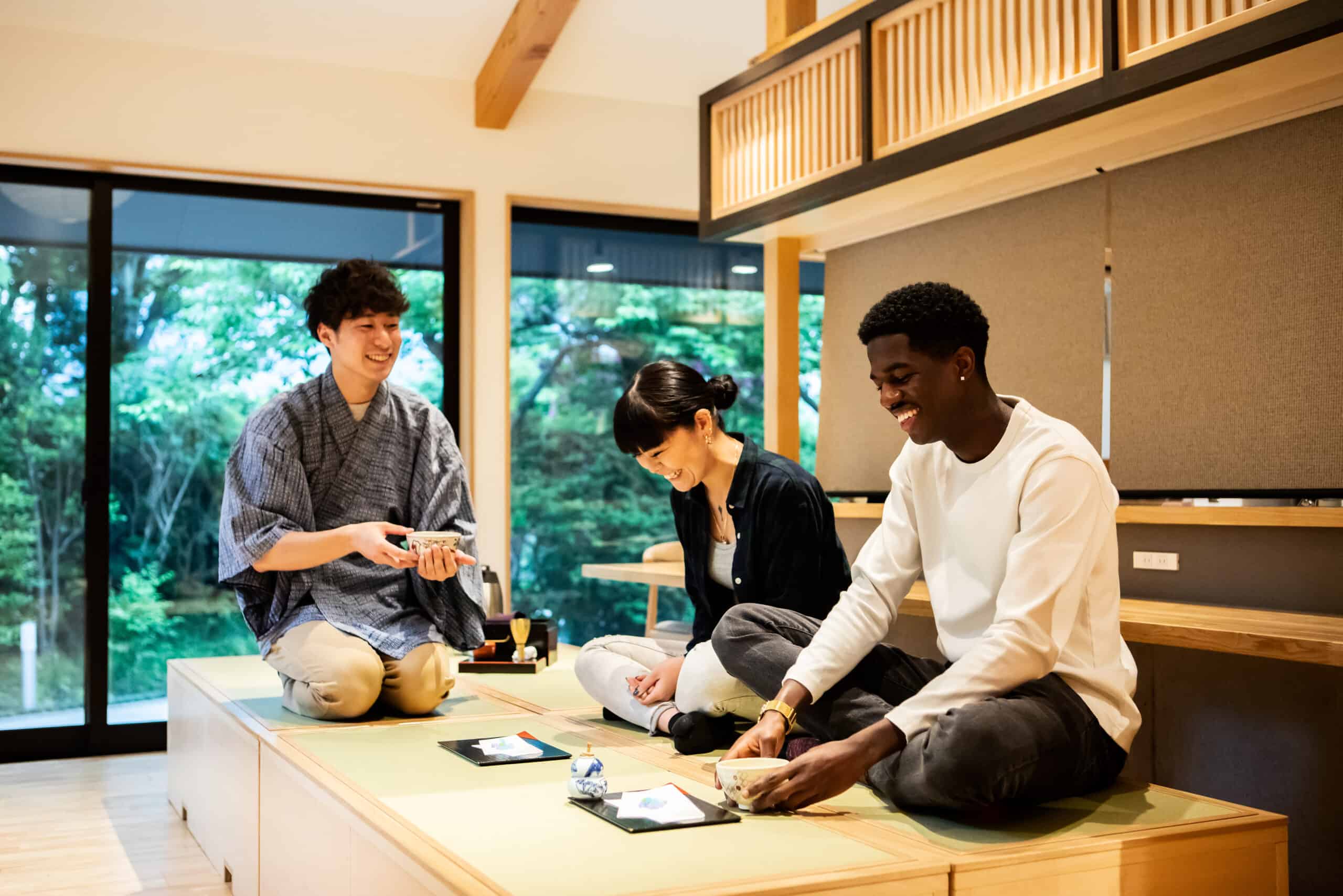 Three people of different genders and ethnicities enjoying tea and conversation in a traditional Japanese-style room with large windows and a wooden interior.