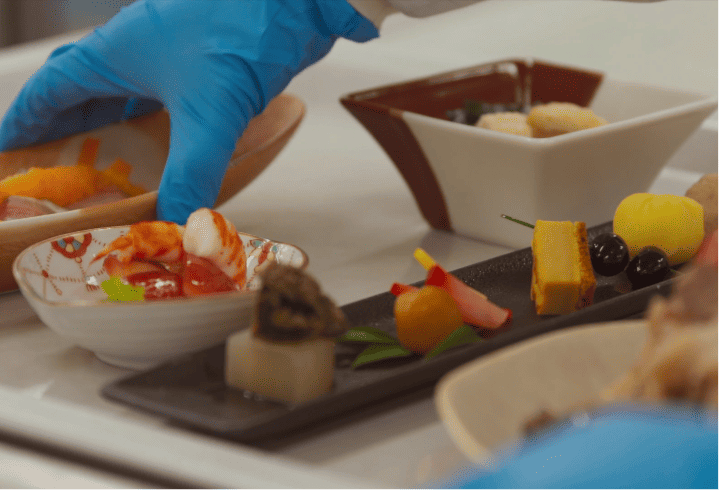 An array of Japanese dishes made with fresh ingredients is served on various dishes, with a person wearing blue gloves placing food.