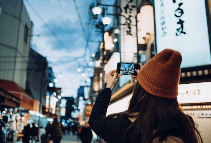 Woman in a beanie taking a photo of a bustling city street at night with her smartphone.