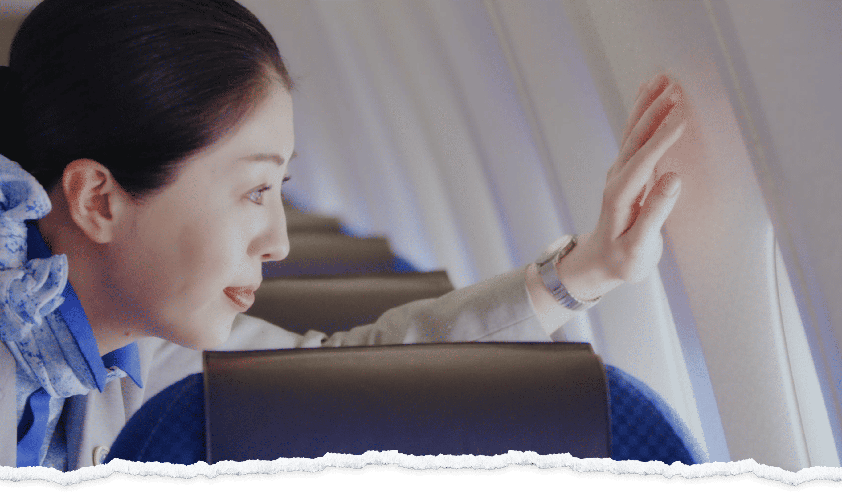 A woman on a plane gently touches the window while looking outside, wearing a watch, blue shirt and a tan jacket.