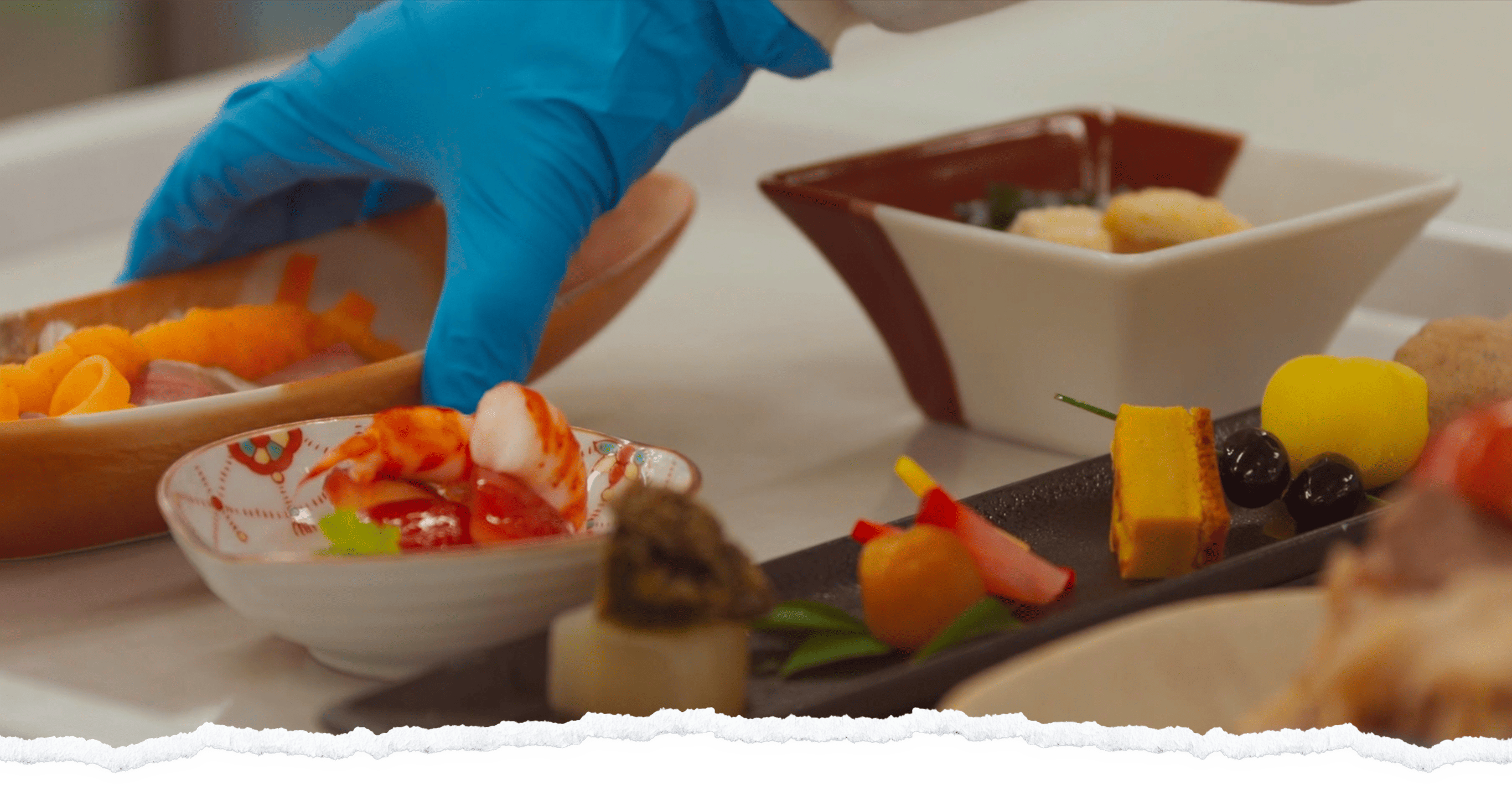 An array of Japanese dishes made with fresh ingredients is served on various dishes, with a person wearing blue gloves placing food.