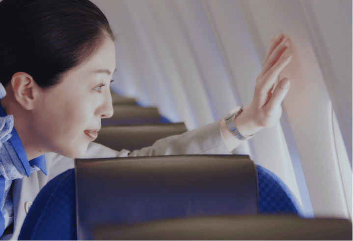 A woman on a plane gently touches the window while looking outside, wearing a watch, blue shirt and a tan jacket.