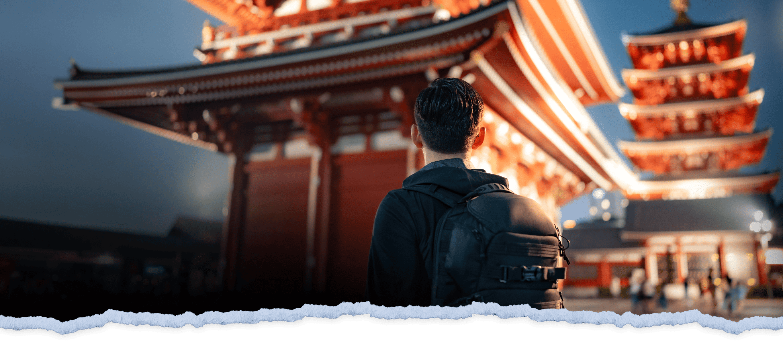 A traveler with a backpack sitting and looking at a traditional Japanese temple with striking red and white structures under a clear sky at dusk.