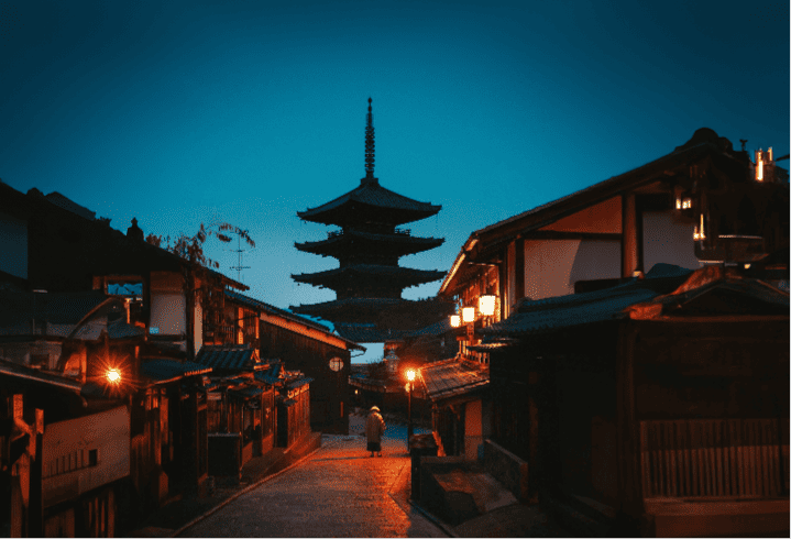 Nighttime view of a traditional Japanese street with a five-tiered pagoda towering in the background and illuminated houses lining the street.