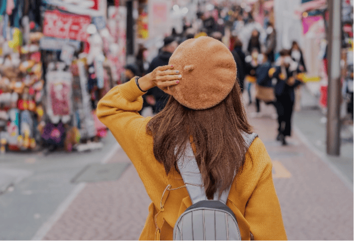 A stylish tourist wearing a backpack holds her beret in place as she walks down a bustling city street with shops in Tokyo.