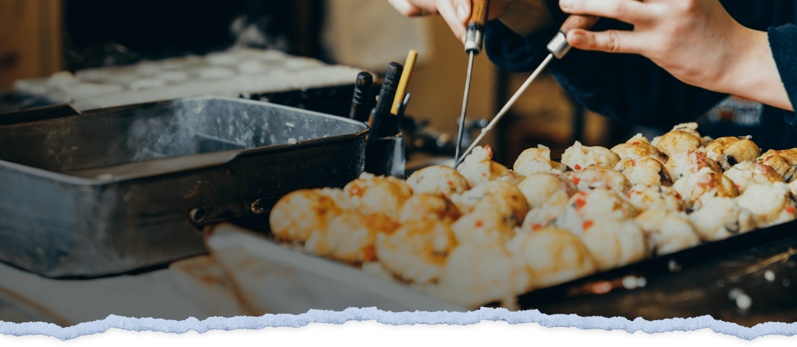 A hand holds utensils that place takoyaki onto a serving plate.