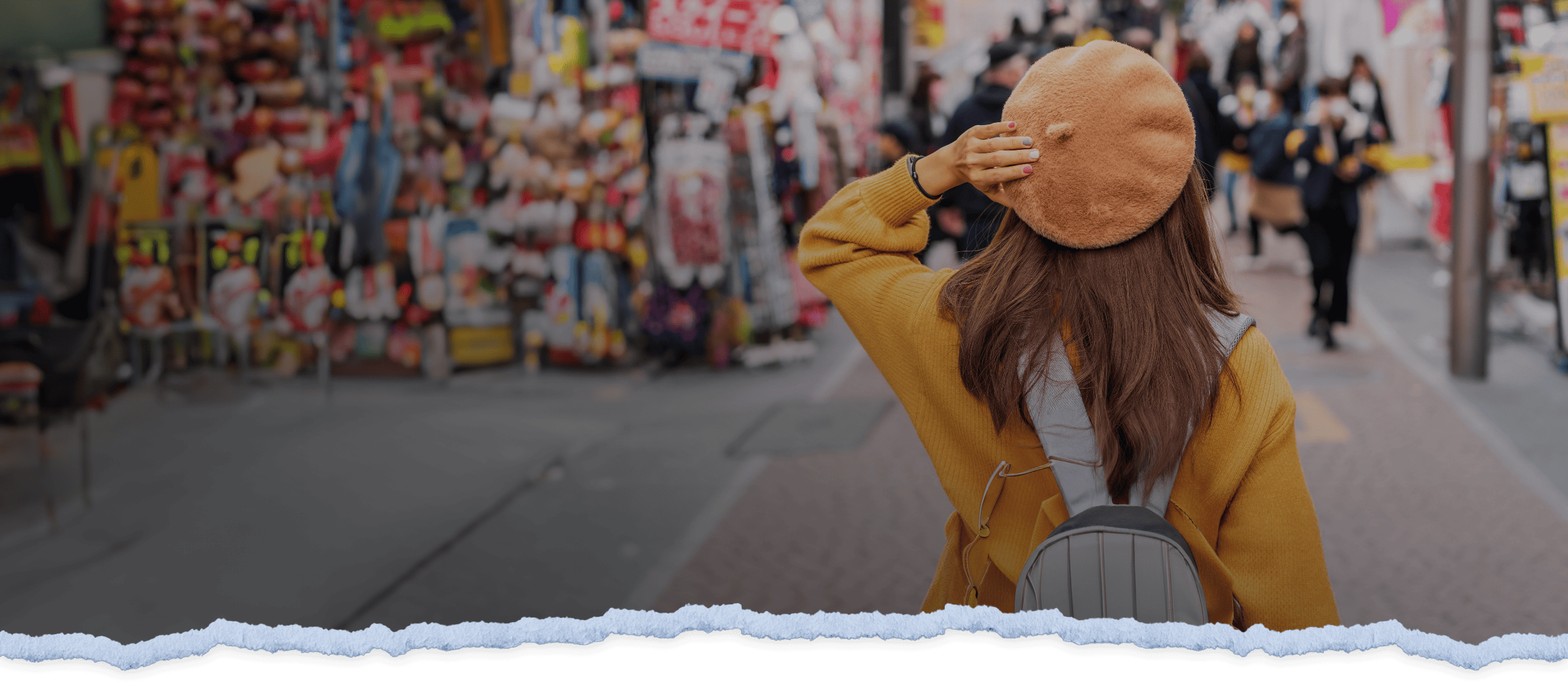 A stylish tourist wearing a backpack holds her beret in place as she walks down a bustling city street with shops in Tokyo.