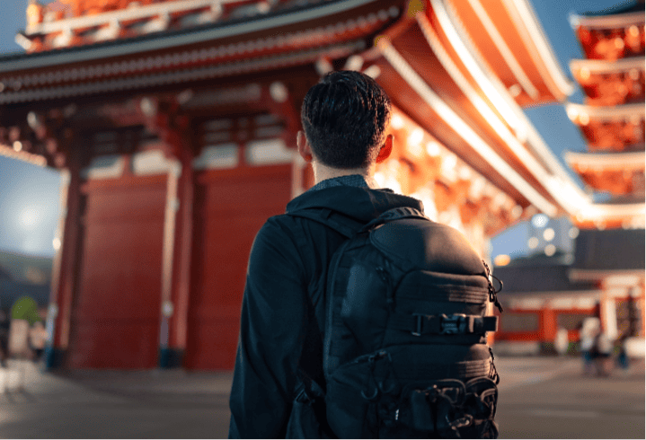A traveler with a backpack sitting and looking at a traditional Japanese temple with striking red and white structures under a clear sky at dusk.
