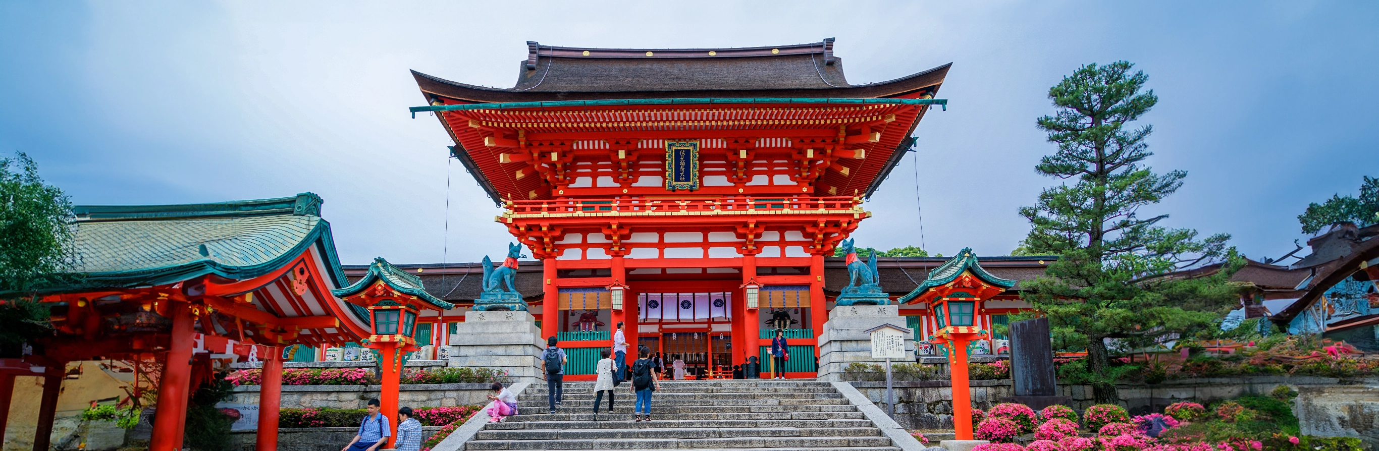 Panoramic view of a vibrant red Japanese temple gate with steps leading up to it, flanked by lush greenery under a cloudy sky, with visitors approaching the entrance.