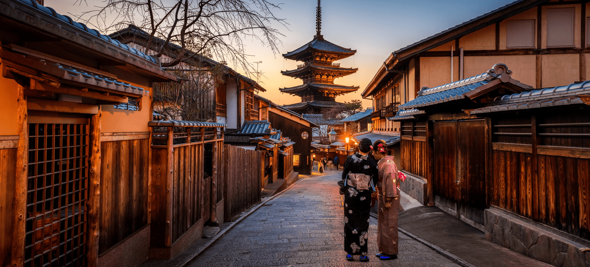b)	Two women wearing kimonos hold their heads close in conversation on a historic Japanese street with a pagoda in the foreground at sunset.