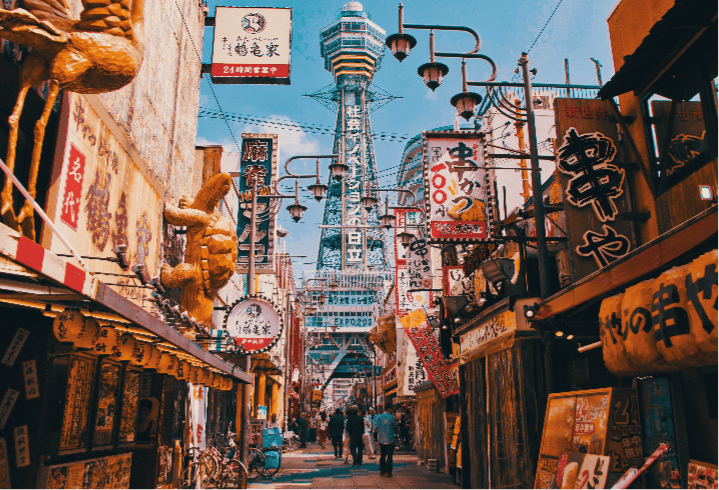 A vibrant street scene in Osaka, Japan, with various traditional signs and lanterns, including a striking blue tower visible in the background, set under a clear sky.