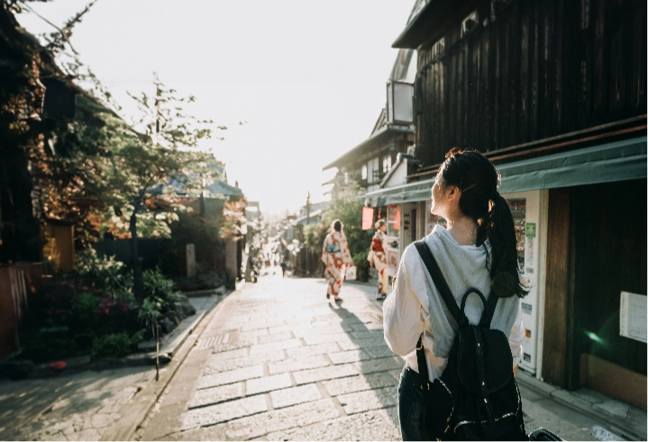 A woman wearing a backpack smiles in the sun on a Japanese street with a suitcase at her side.
