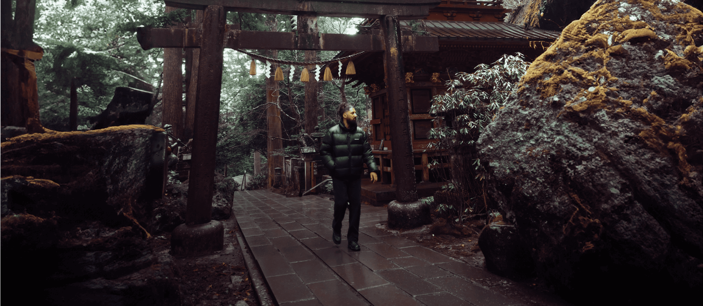 A man in a black jacket walks under the gate of a historic Japanese temple on an overcast day.
